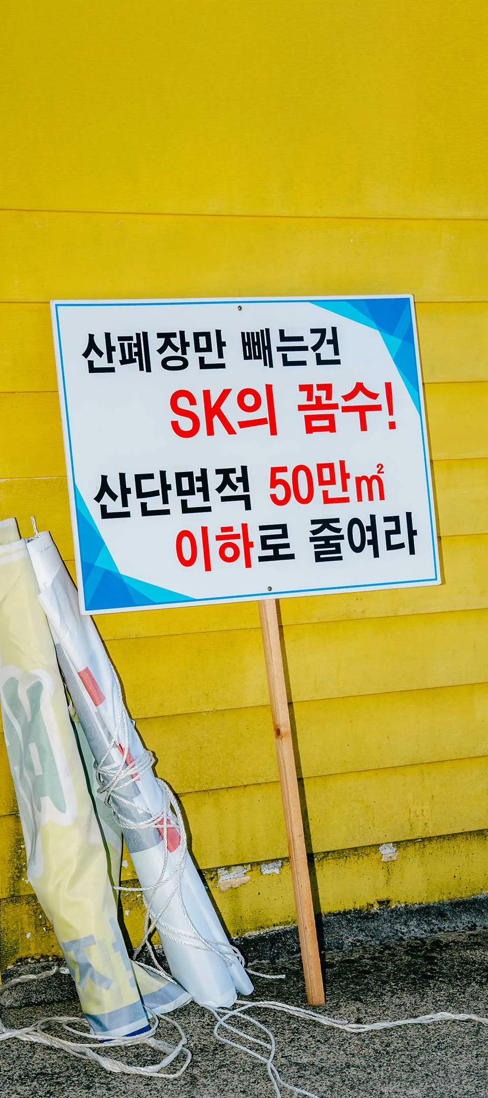 산폐장만 빼는 건 에스케이의 꼼수 산단면적 50만 제곱미터 이하로 줄여라 라고 쓰여 있는 두 사람의 투쟁용 피켓이 조순희의 집 노란색 외벽에 세워져 있는 사진.