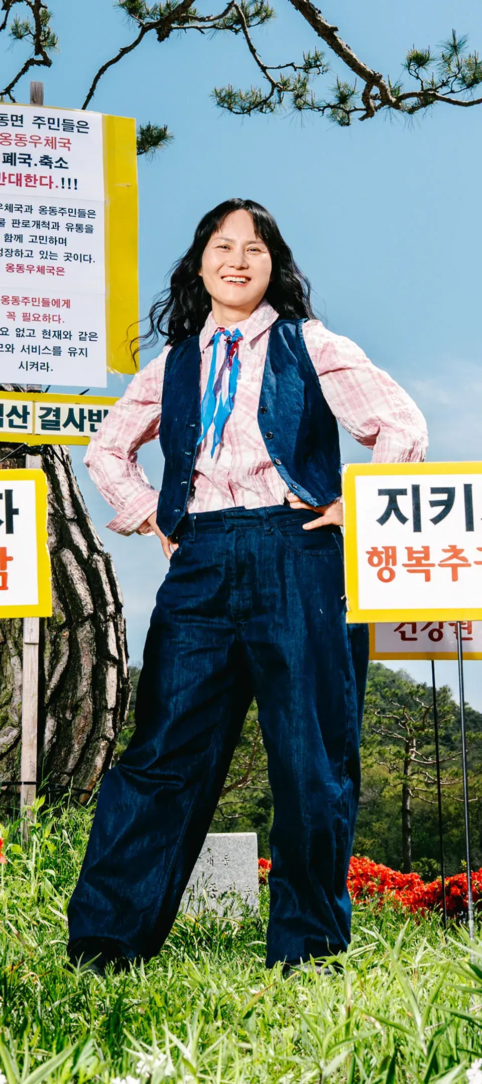 석산 결사반대, 지키자 행복추구권 등의 피켓들이 세워진 옹동면사무소 앞 소나무 조경지를 배경으로 데님 소재의 캐주얼한 탐정 복장을 입은 엄성자가 양손을 허리에 얹고 자신감 있는 미소로 정면을 바라보는 사진.