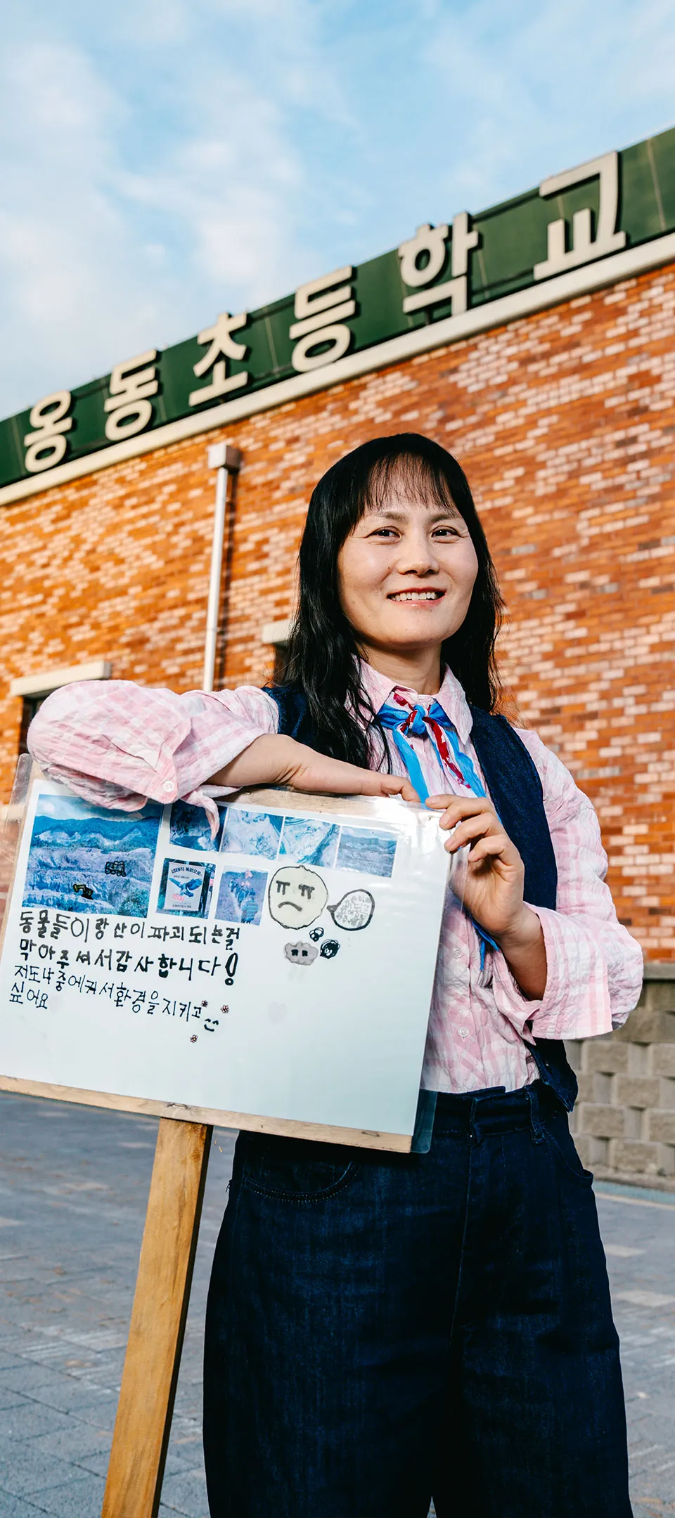 옹동초등학교 학생이 제작한 피켓에 오른팔을 괸 채, 따뜻하고 인자한 미소를 지으며 정면을 바라보는 엄성자의 사진. 피켓에는 동물들이랑 산이 파괴되는 걸 막아주셔서 감사합니다! 저도 나중에 커서 환경을 지키고 싶어요.라고 쓰여 있다.